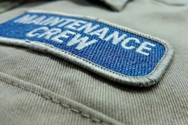 Close-up of a blue and white embroidered Maintenance Crew patch on a tan jacket showing thread fraying and surface fuzz.