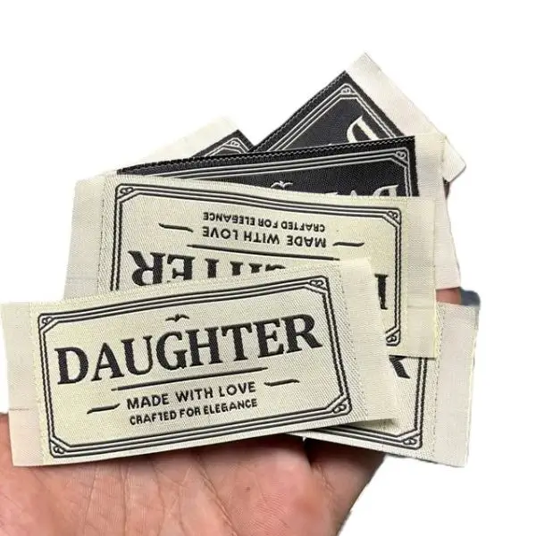 Stack of cream custom woven hem tags held in hand, showcasing sharp, legible serif text perfect for premium apparel lines.