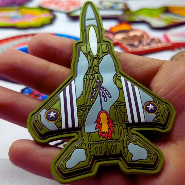 A hand holding a military-style fighter jet custom PVC patch with intricate 3D molding and olive green tones.