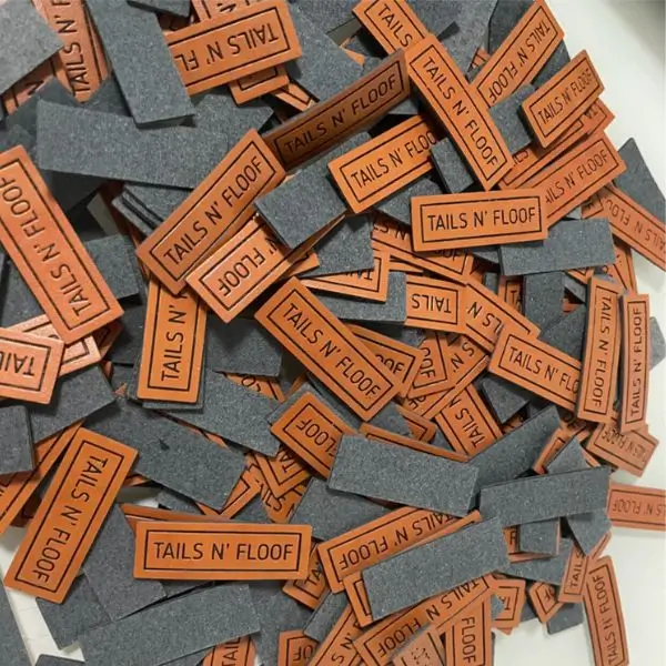 A bulk pile of small rectangular leather labels for Tails N' Floof showing grey felt backing